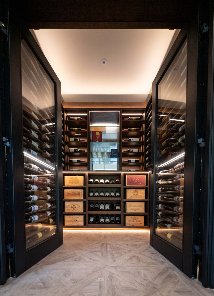 Double doors opening out to show the wine cellar inside. Dark wood wine racks show off the bottles and lighting can be seen, shining down on the wine bottle labels.