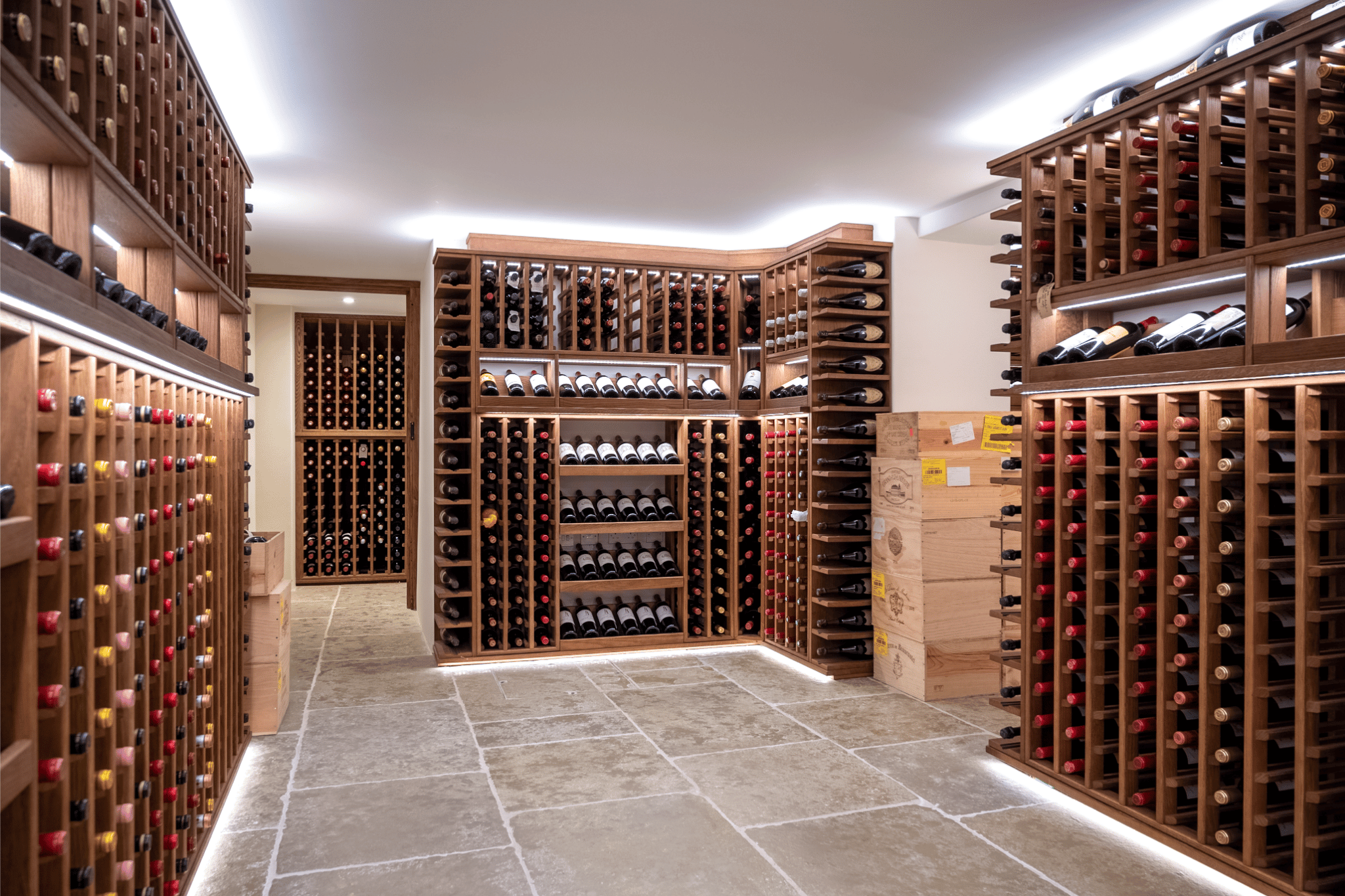 A dark oak wine cellar. The main room has bright white walls, of which wine racking sits against. The wine racking is dark in colour. Beyond the room, a further wine rack can be seen through the open door, against the back wall of the room behind.